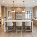 Bright luxury kitchen remodel in Dallas featuring a large white waterfall island, light wood cabinets, wide plank wood flooring, and stainless steel appliances.